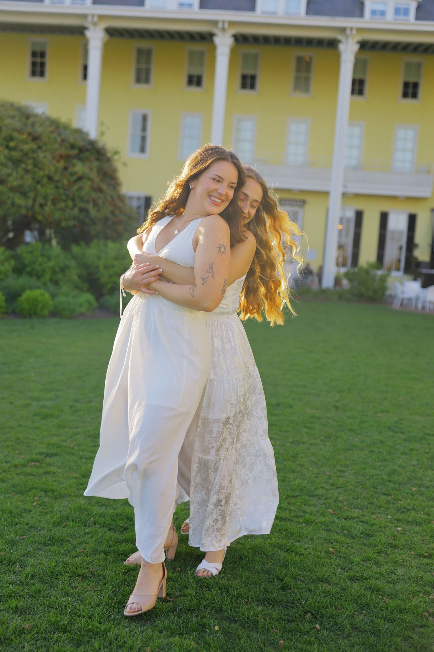 Two women in white dresses embrace on green lawn before a yellow Victorian building in golden hour light.
