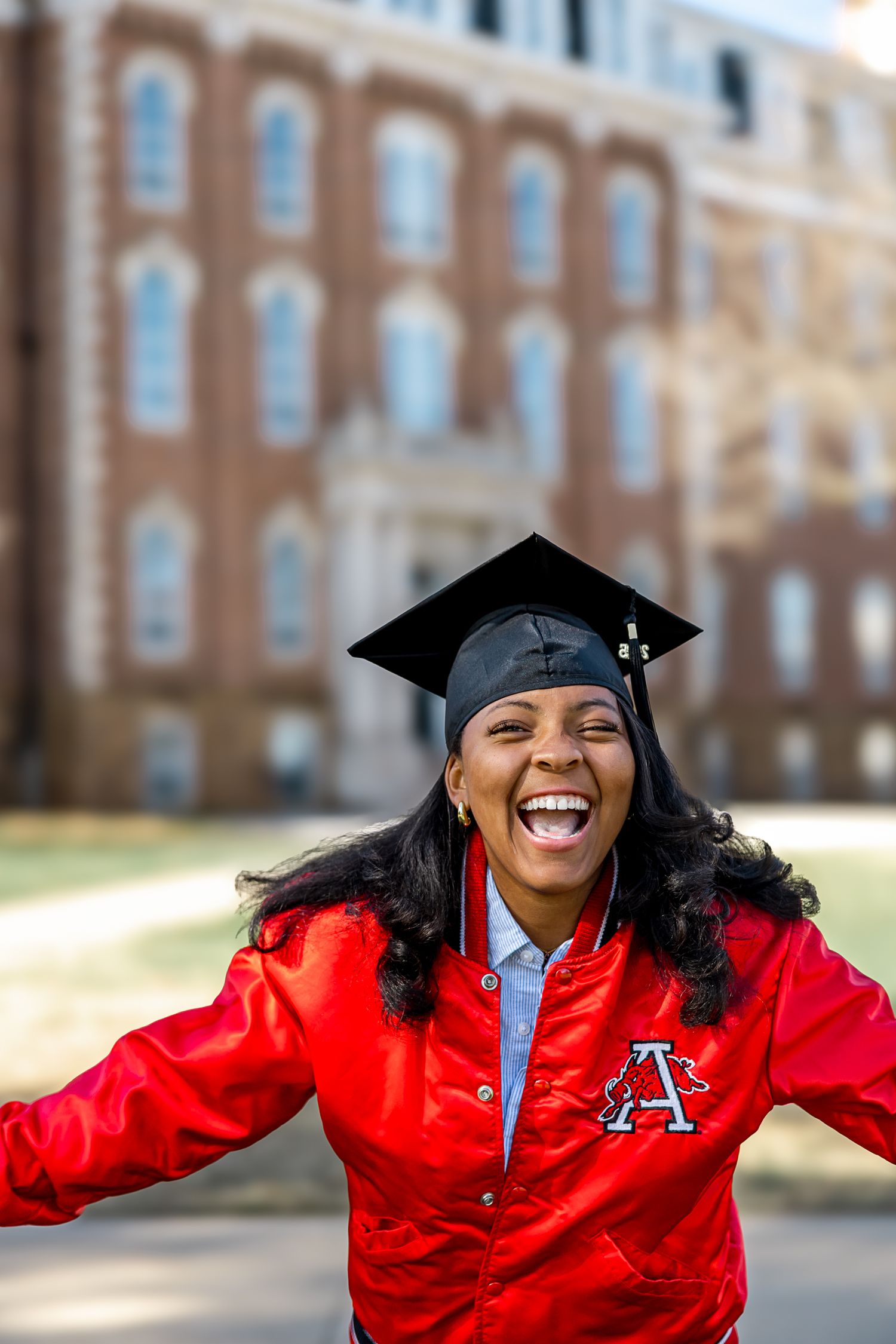 Graduate in red academic regalia and cap smiles joyfully while posing outdoors on campus during commencement.
