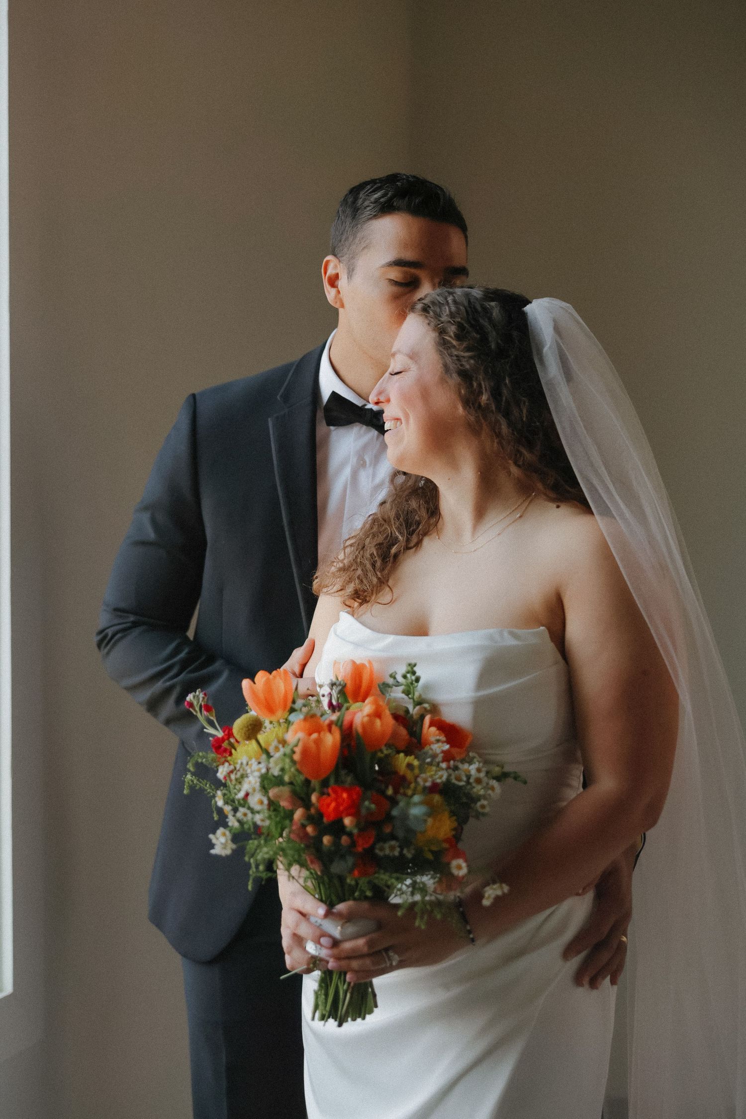 Wedding couple poses indoors with a vibrant orange and red floral bouquet.