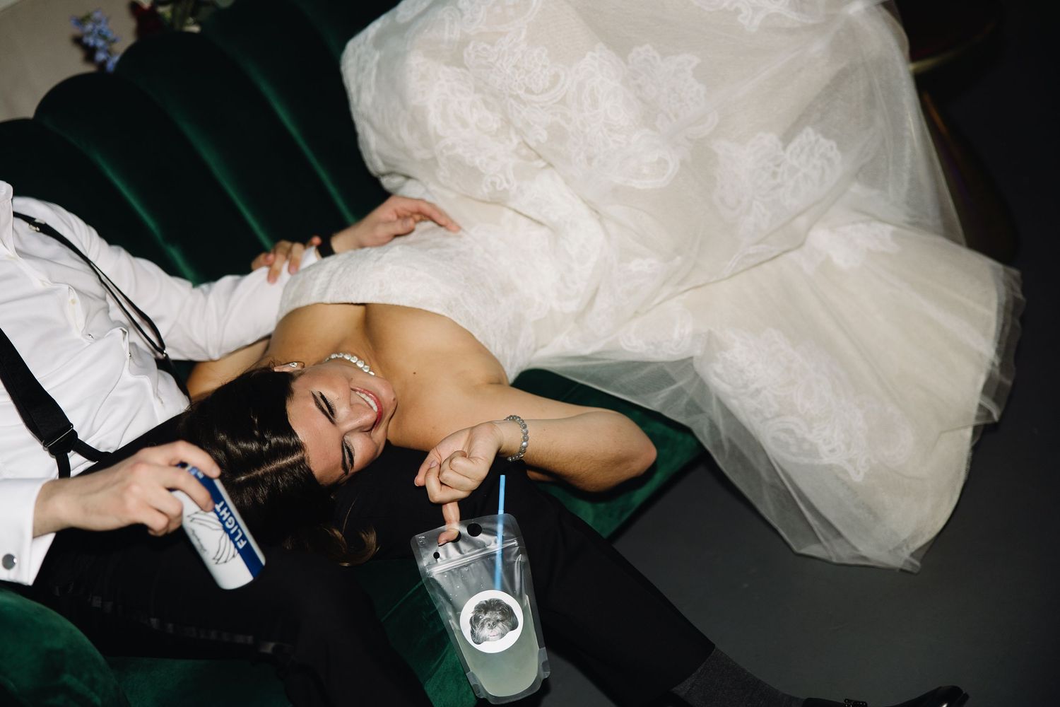 Wedding dress flowing on green velvet couch with drinks in a candid party moment.