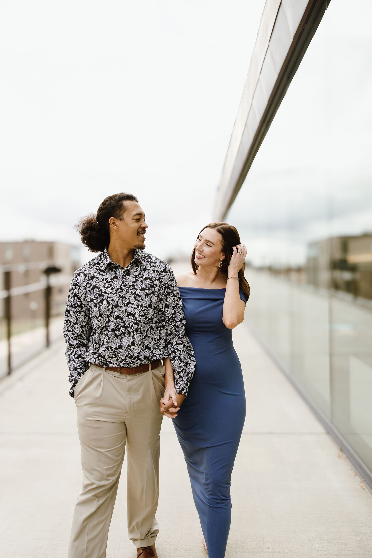 A couple holds hands and smiles at each other while walking along a modern glass corridor outdoors.
