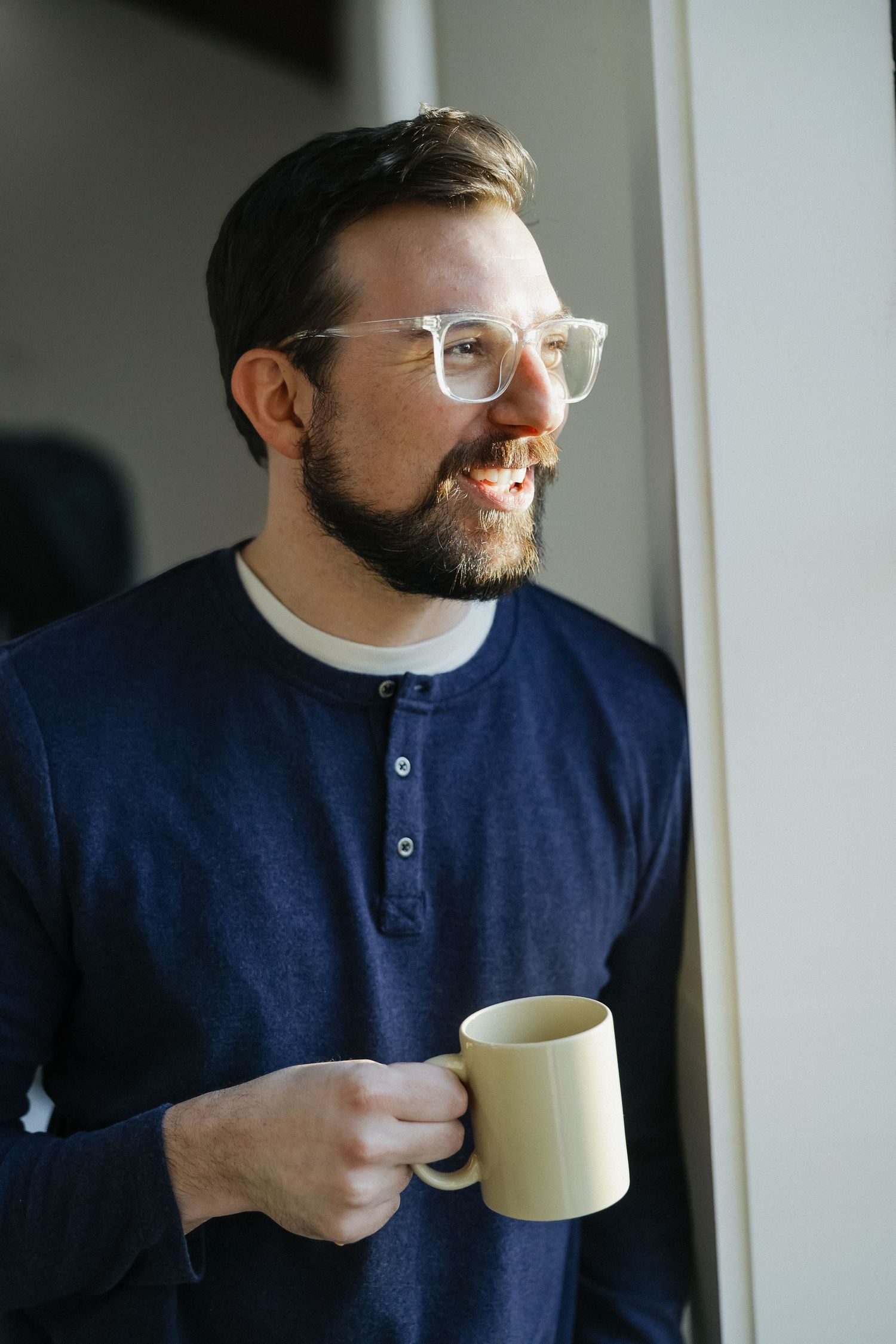 Smiling bearded man with glasses holding a yellow mug, wearing a navy henley shirt, standing by a window.