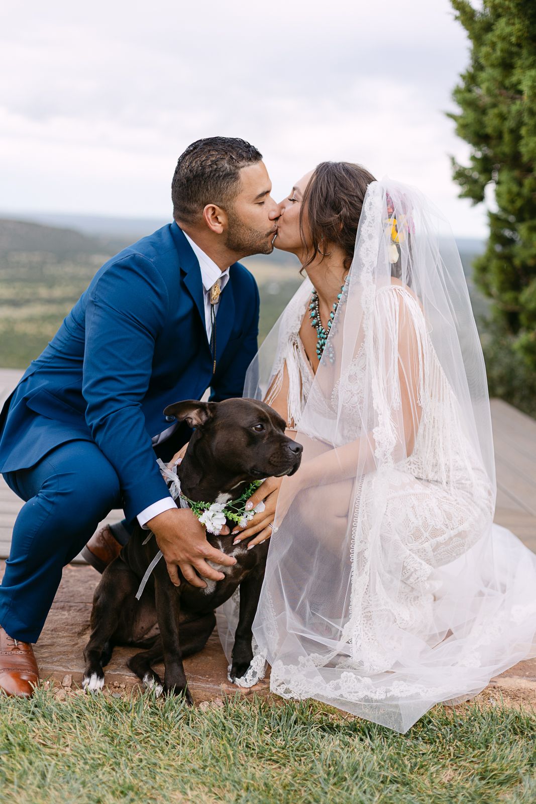 Couple sharing romantic kiss with their black dog during outdoor wedding photoshoot.