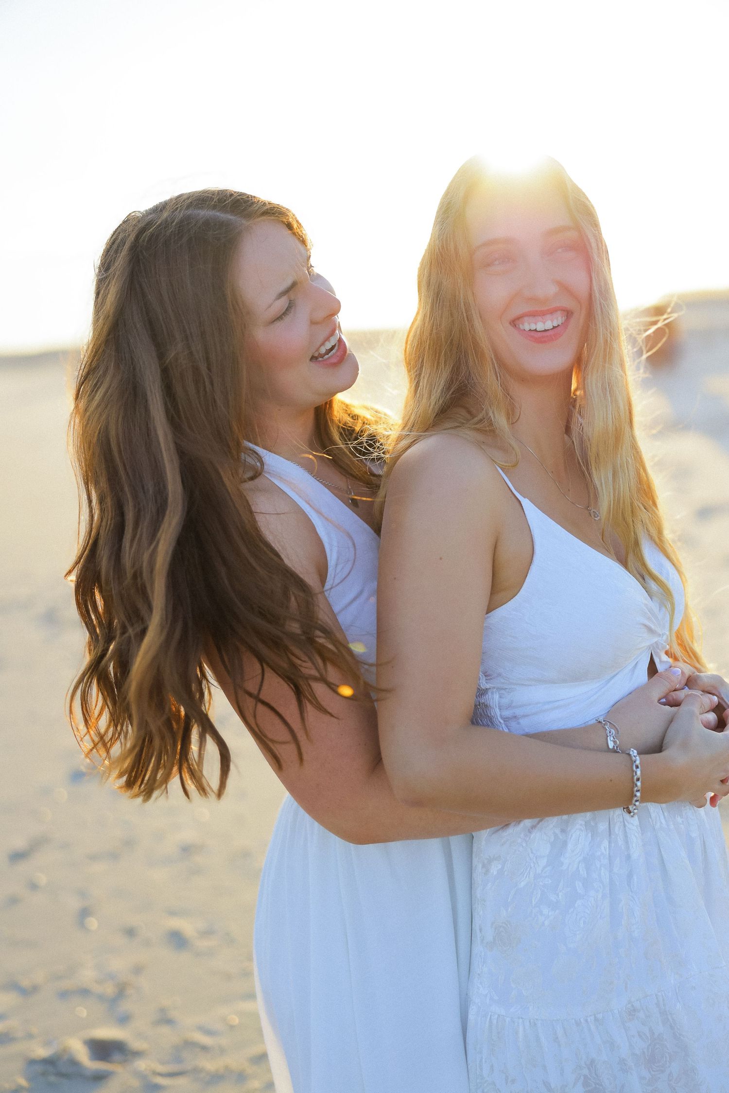 Two young women in white dresses laughing and embracing on a sunny beach at golden hour.