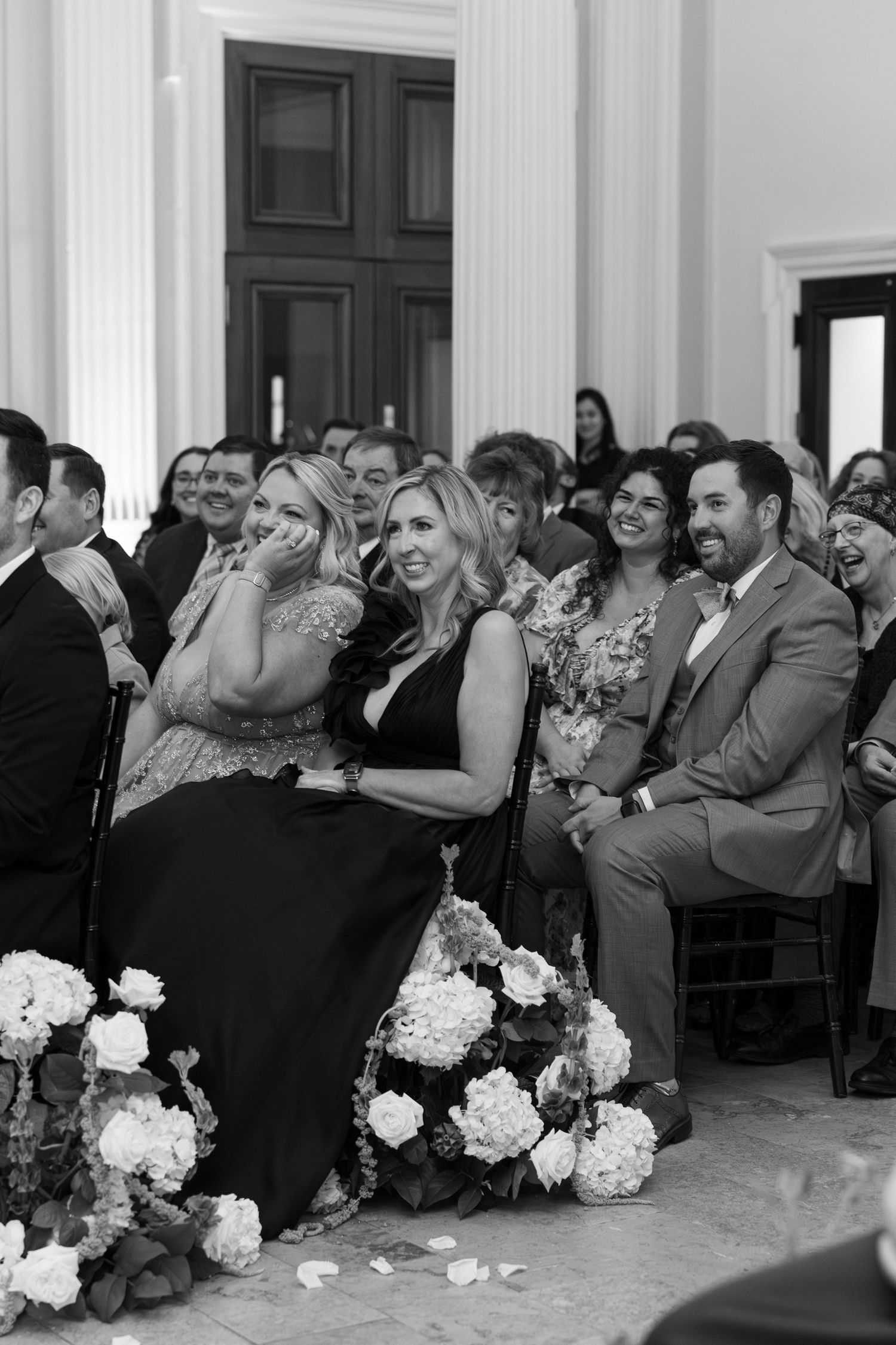 Black and white photo of wedding guests laughing and crying with joy during ceremony, floral arrangements in foreground.