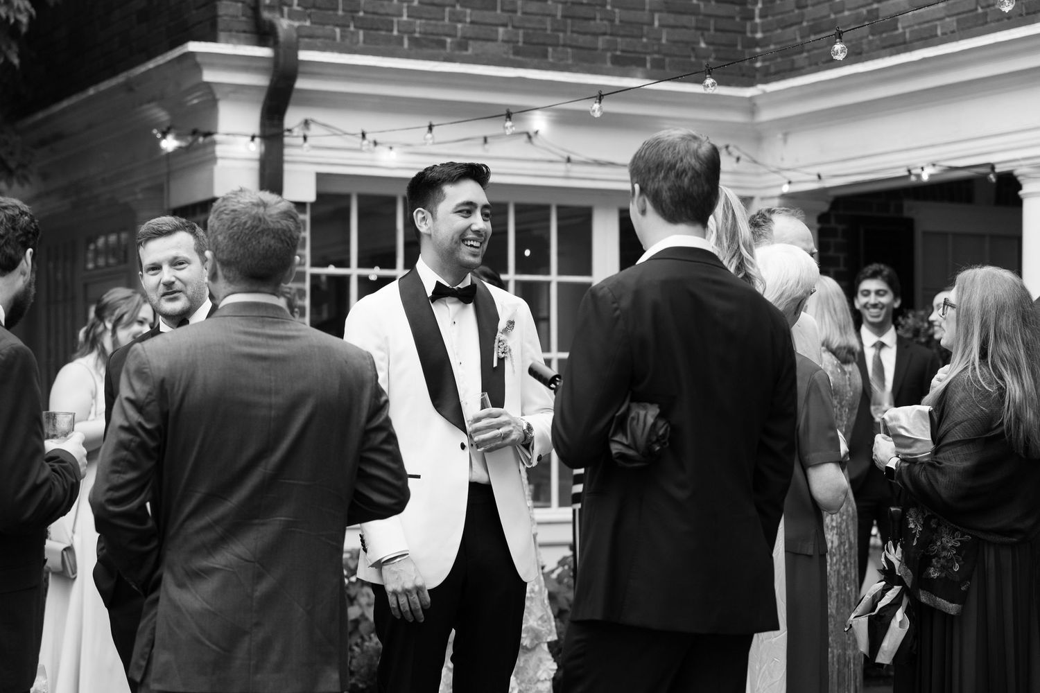 Black and white photo of guests mingling outdoors at a wedding reception, groom in white tuxedo jacket.