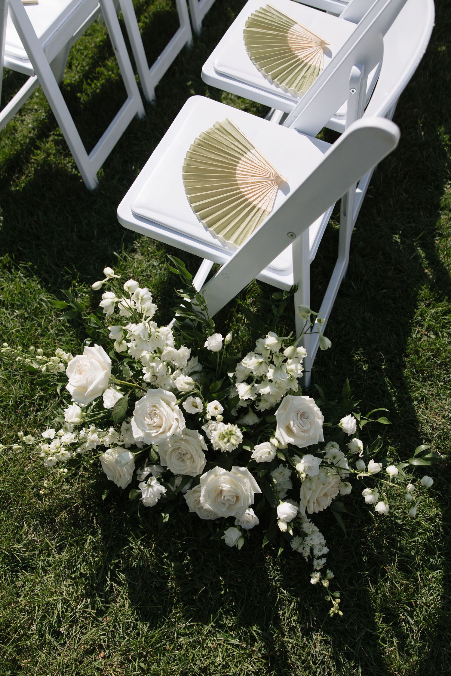 White folding chairs with gold fans at an outdoor wedding ceremony, accented by elegant white rose floral arrangements on green grass.