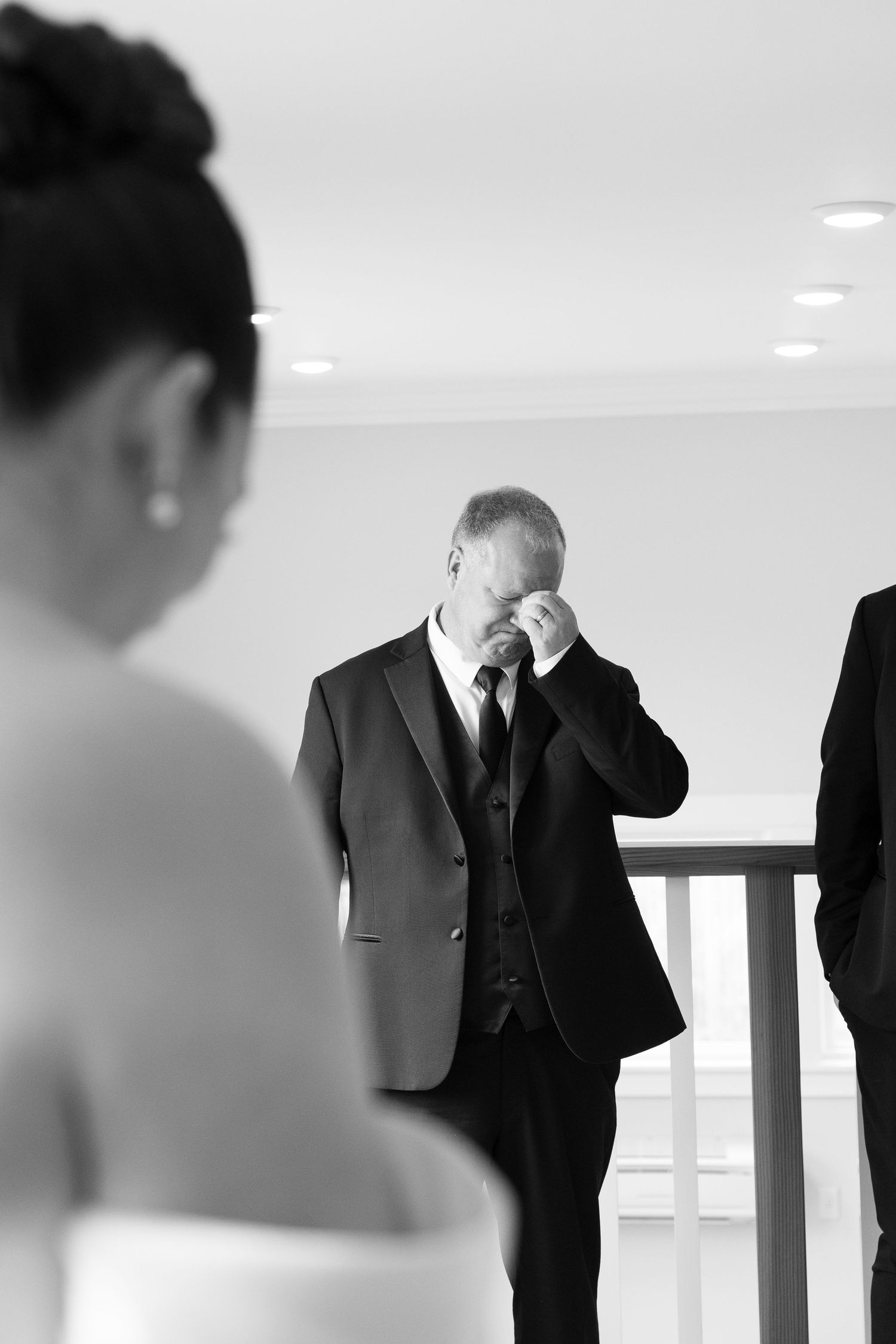 Black and white photo of emotional groom wiping tears as bride approaches during wedding ceremony.
