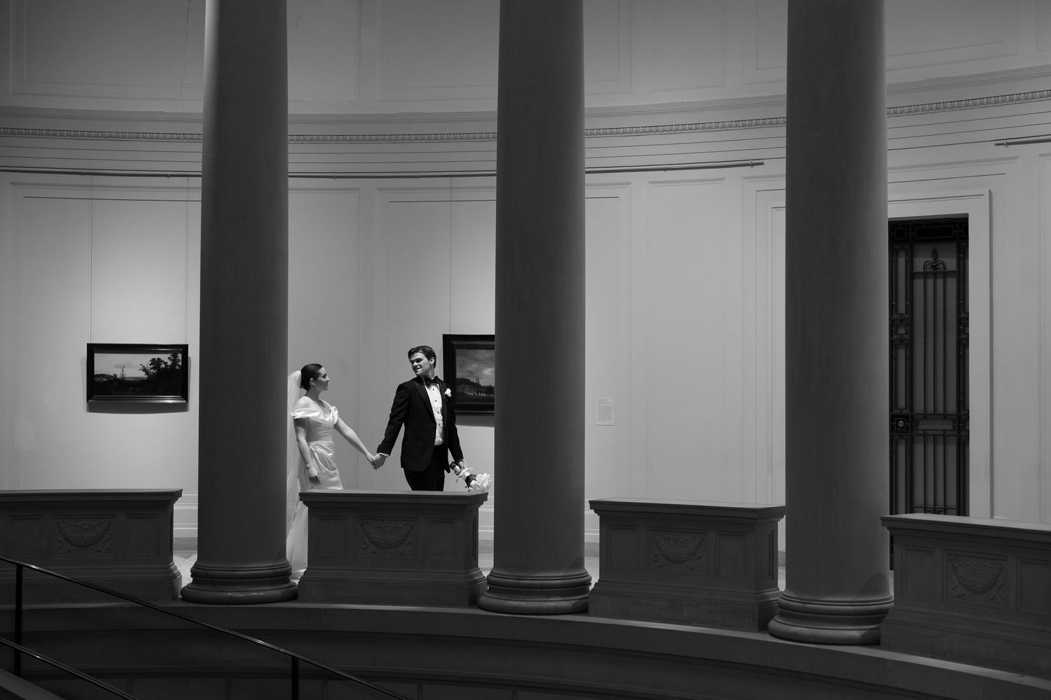 Black and white photo of a bride and groom holding hands between grand columns in an elegant museum hall.