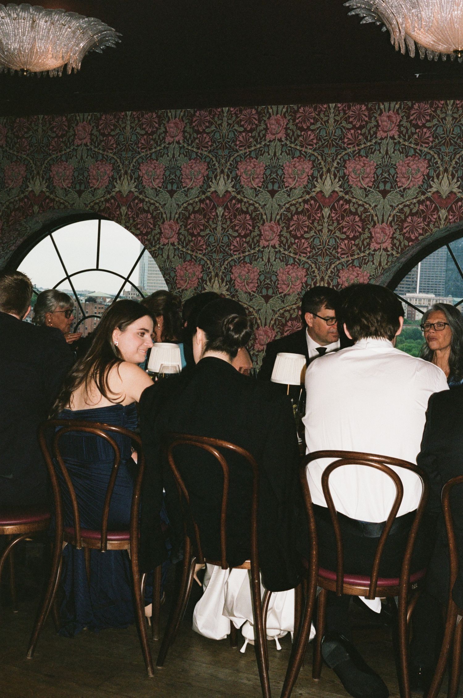 Elegantly dressed guests seated at round tables in a dimly lit venue with ornate floral wallpaper during a formal event.