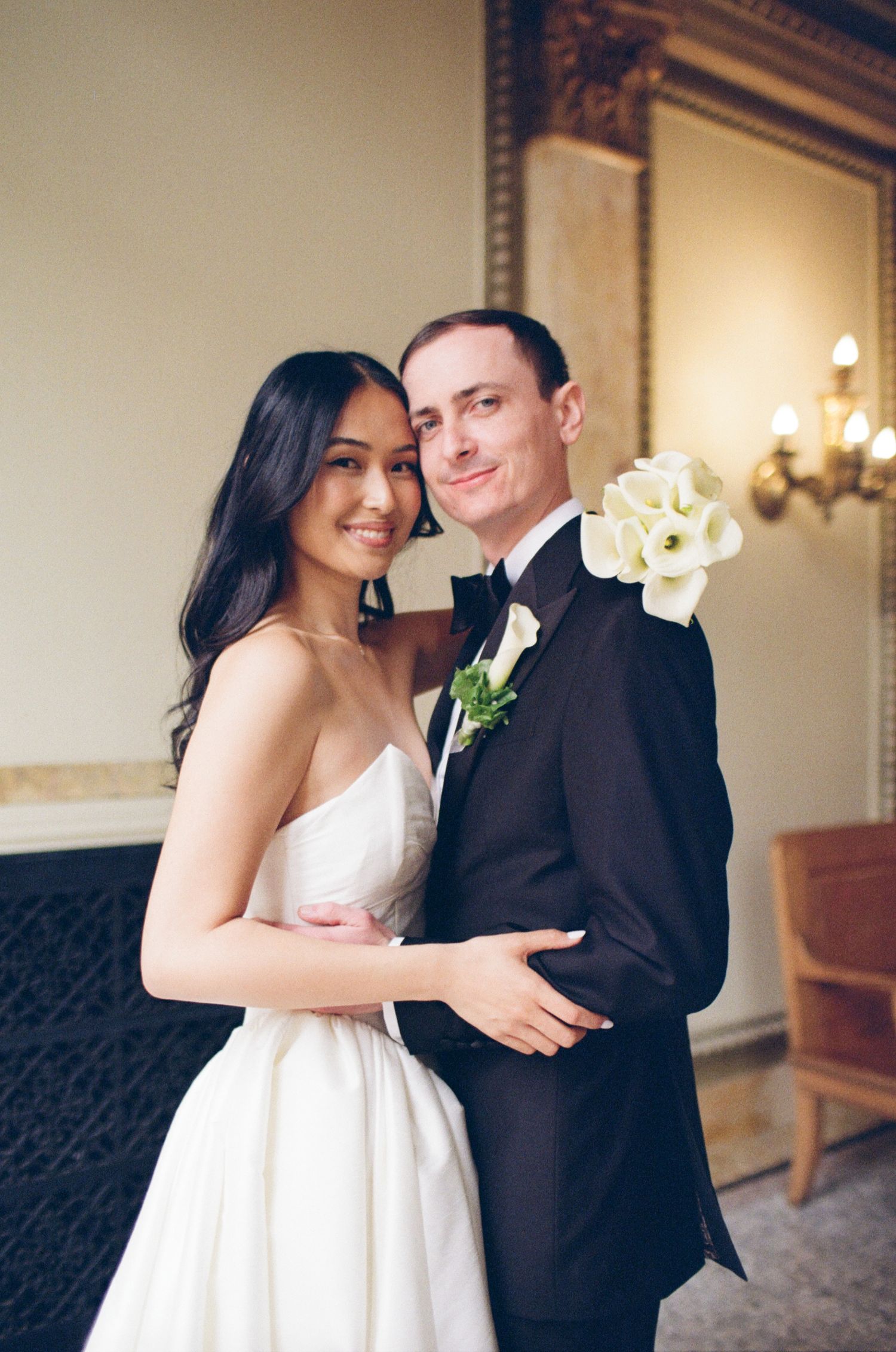 A smiling bride in a white gown embraces a groom in a dark tuxedo with white calla lily boutonniere.