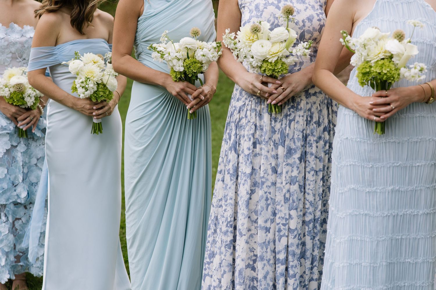 Bridesmaids in mismatched blue floral and pastel gowns holding white and green bouquets at an outdoor wedding.
