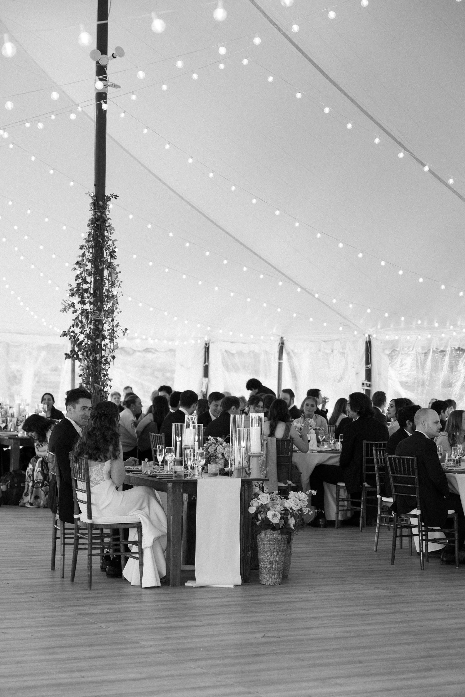 Black and white photo of wedding reception under a tent with string lights, guests seated at tables with floral centerpieces.