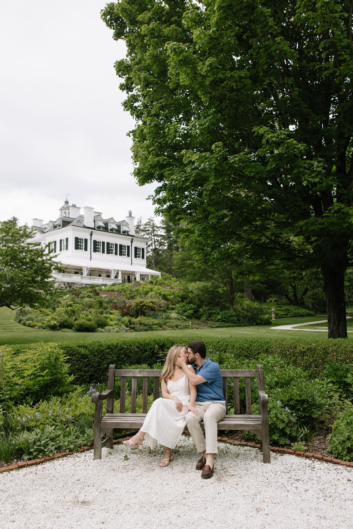 Couple sitting on wooden bench kissing with elegant white New England estate in background.