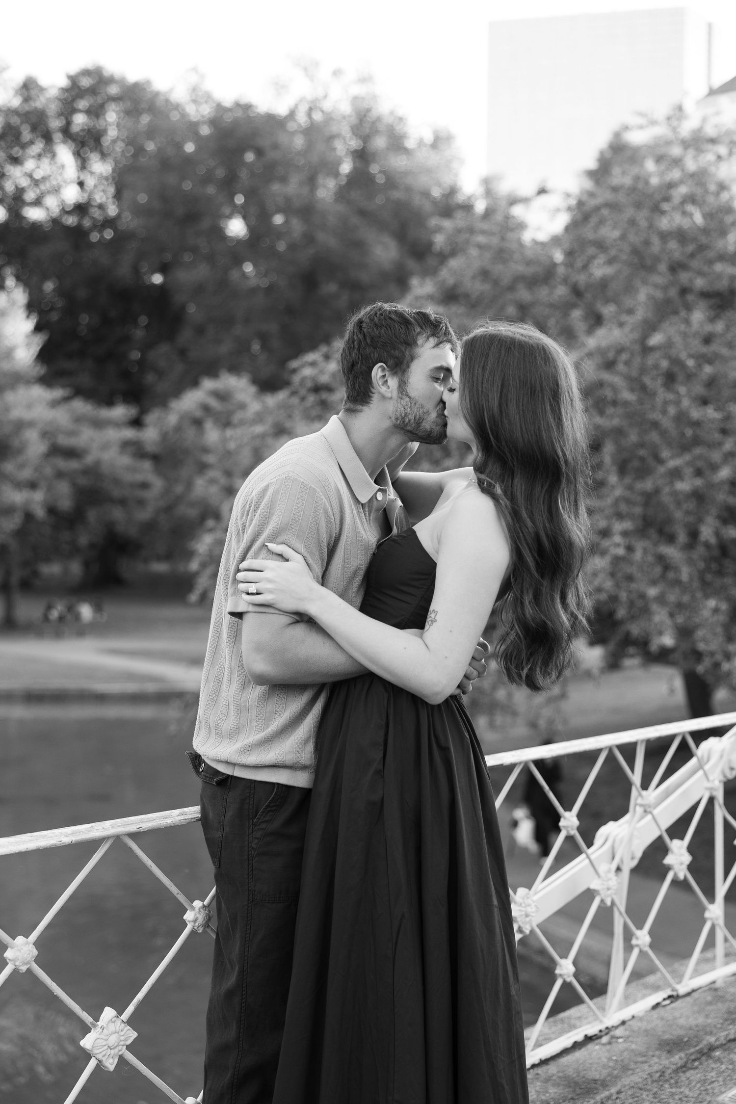 A couple embraces on a white bridge railing with a park landscape behind them.