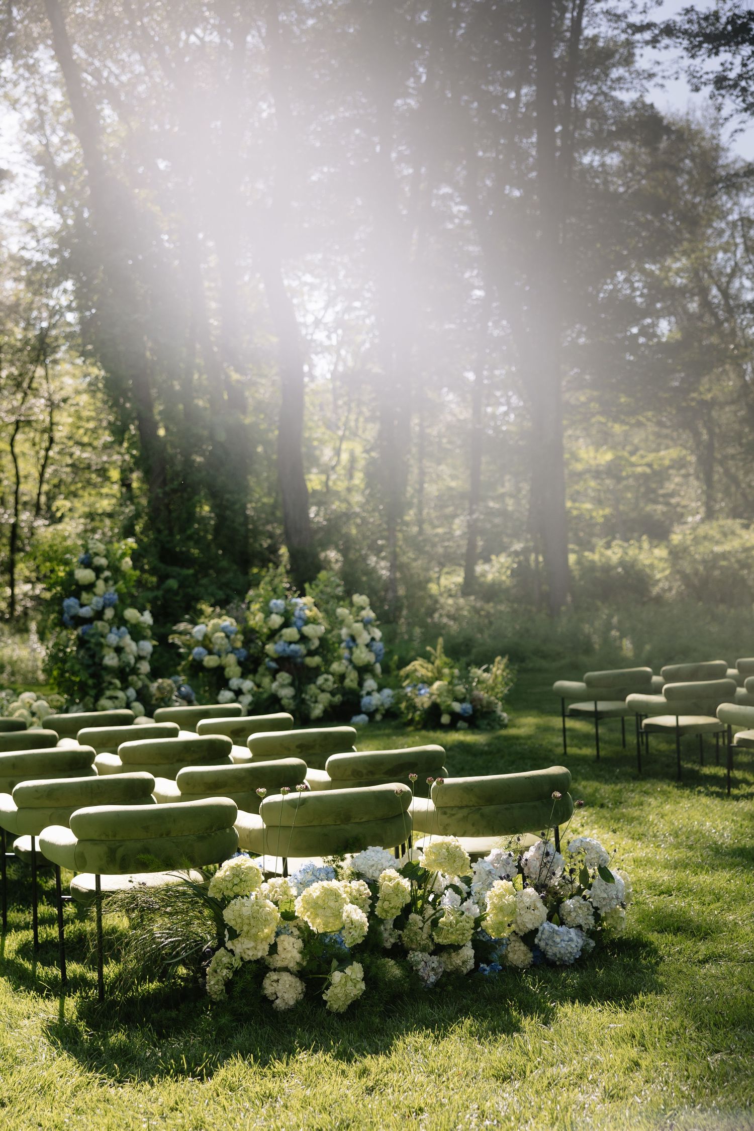 Outdoor wedding ceremony setup with white chairs and floral arrangements in a sunlit wooded area.
