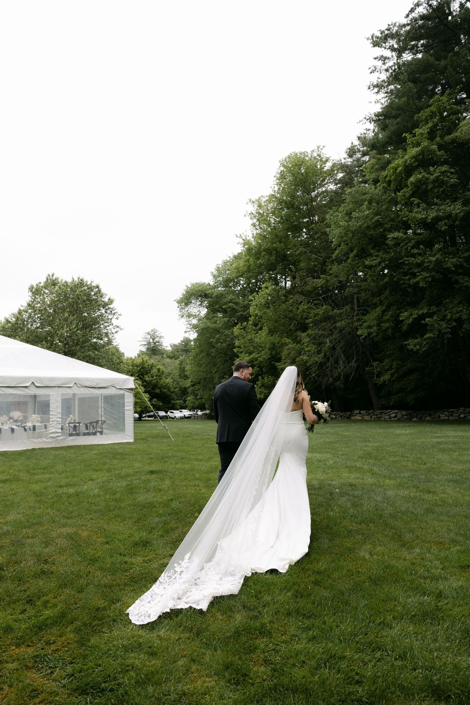 Wedding couple walking across green lawn with flowing white dress and tent in background.