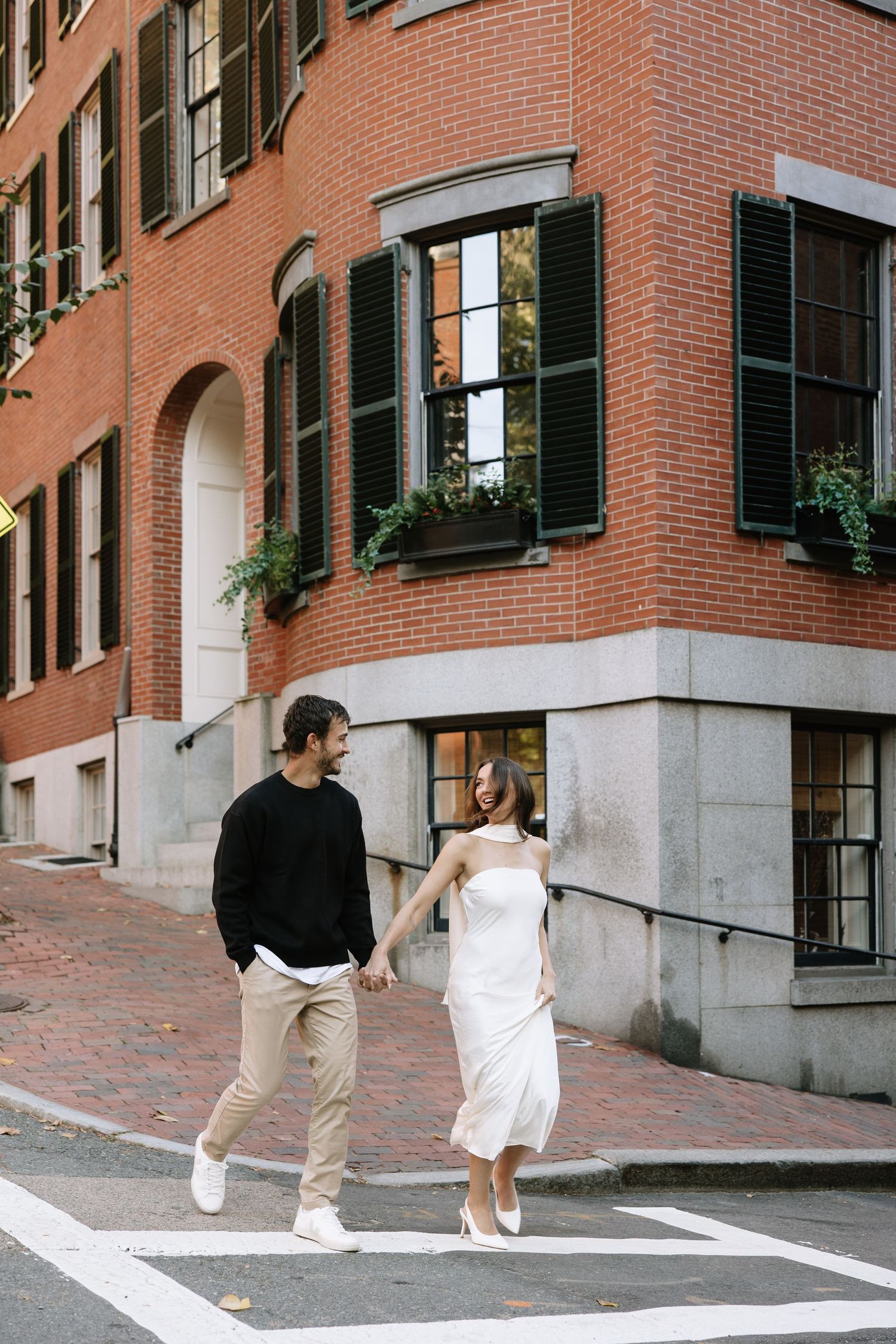 Couple walking together past a red brick building with black shutters and window boxes.