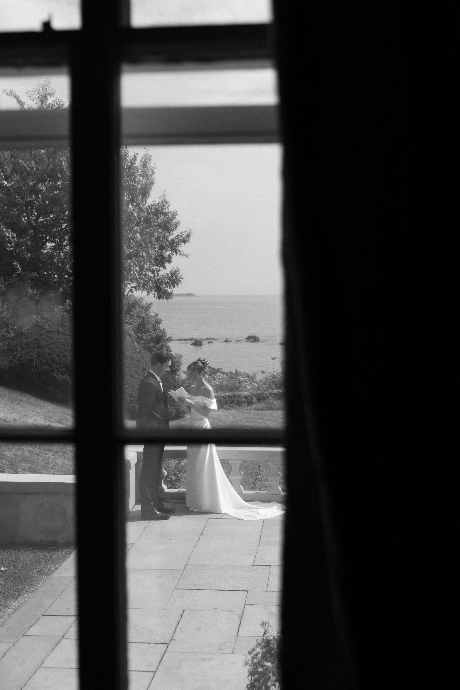 Black and white photo of bride and groom embracing on a seaside terrace, viewed through a window.