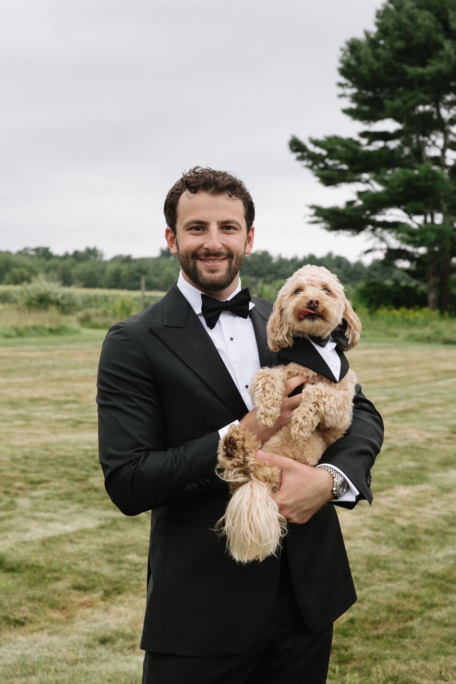 Smiling groom in black tuxedo holding a fluffy golden doodle dog also dressed in a tiny tuxedo outdoors.