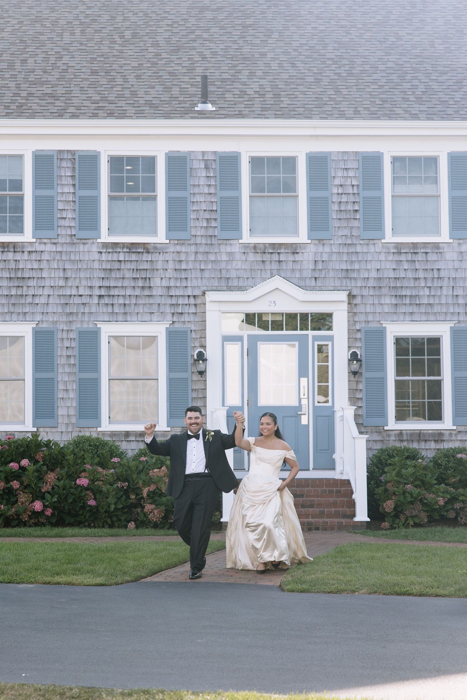Bride and groom celebrate joyfully outside a charming Cape Cod-style venue with blue shutters.