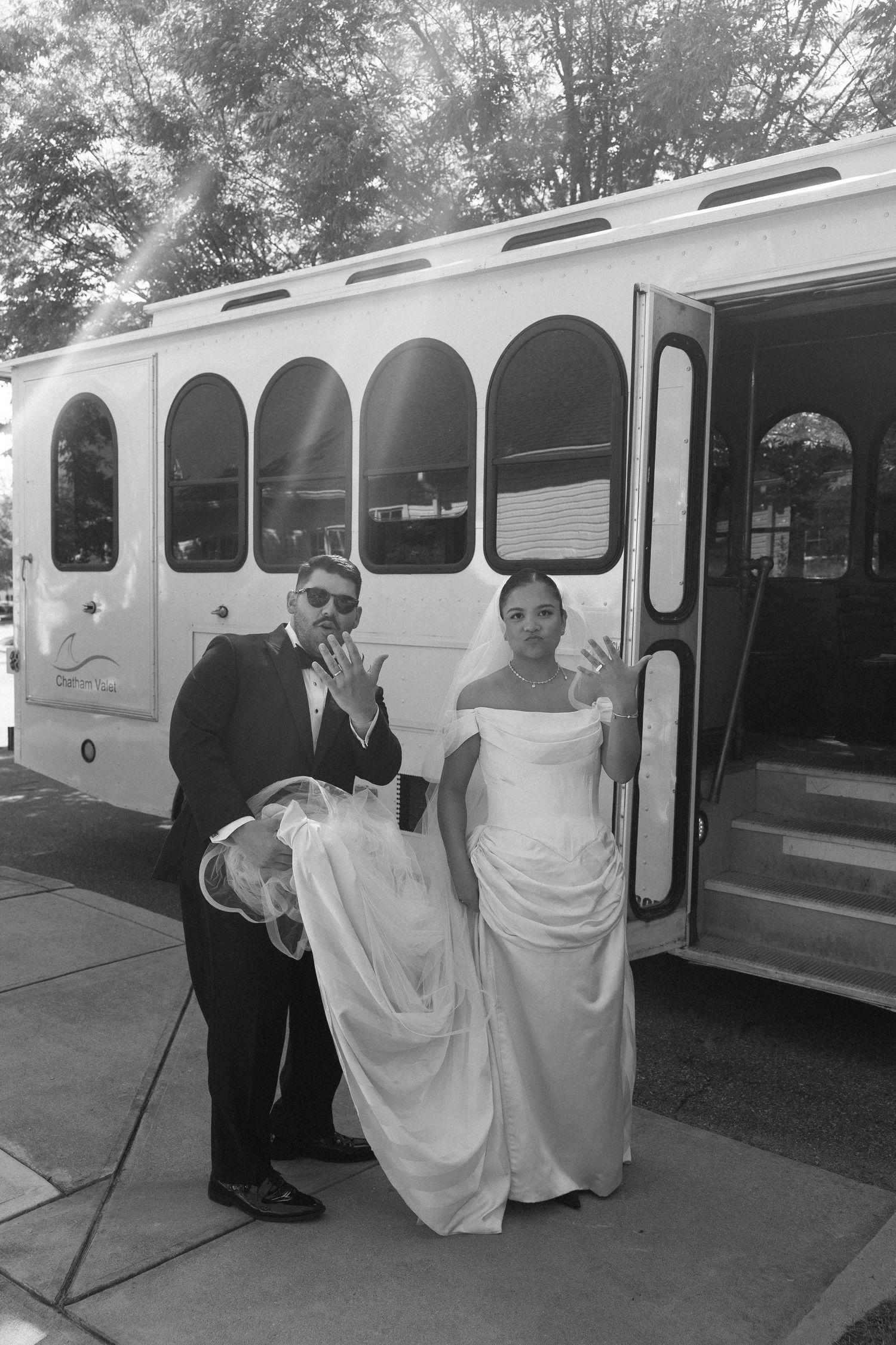 Bride and groom posing in front of a white wedding trolley bus, showing off rings in black and white photo.