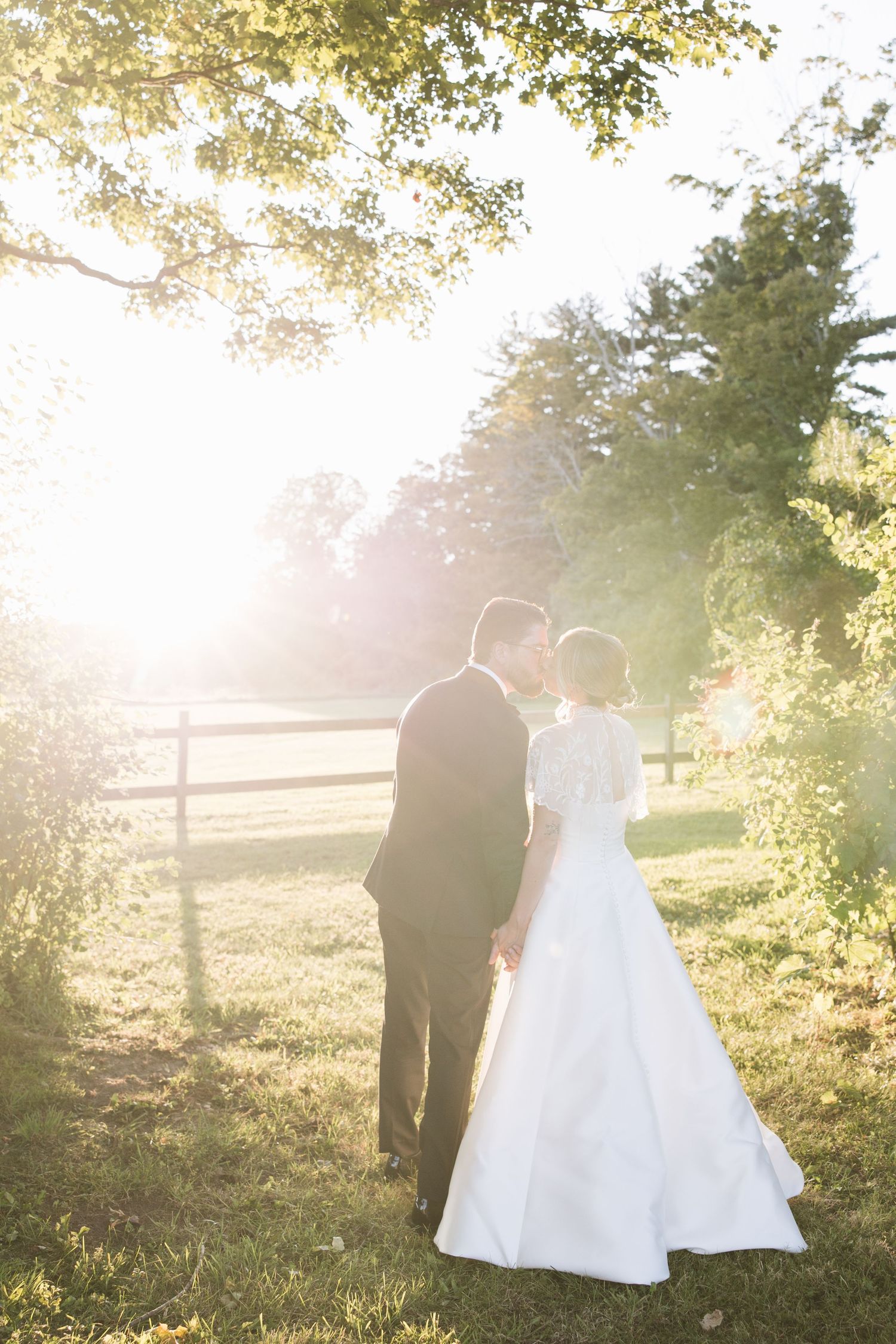 Bride and groom hold hands in golden sunlight among lush green trees at outdoor wedding ceremony.