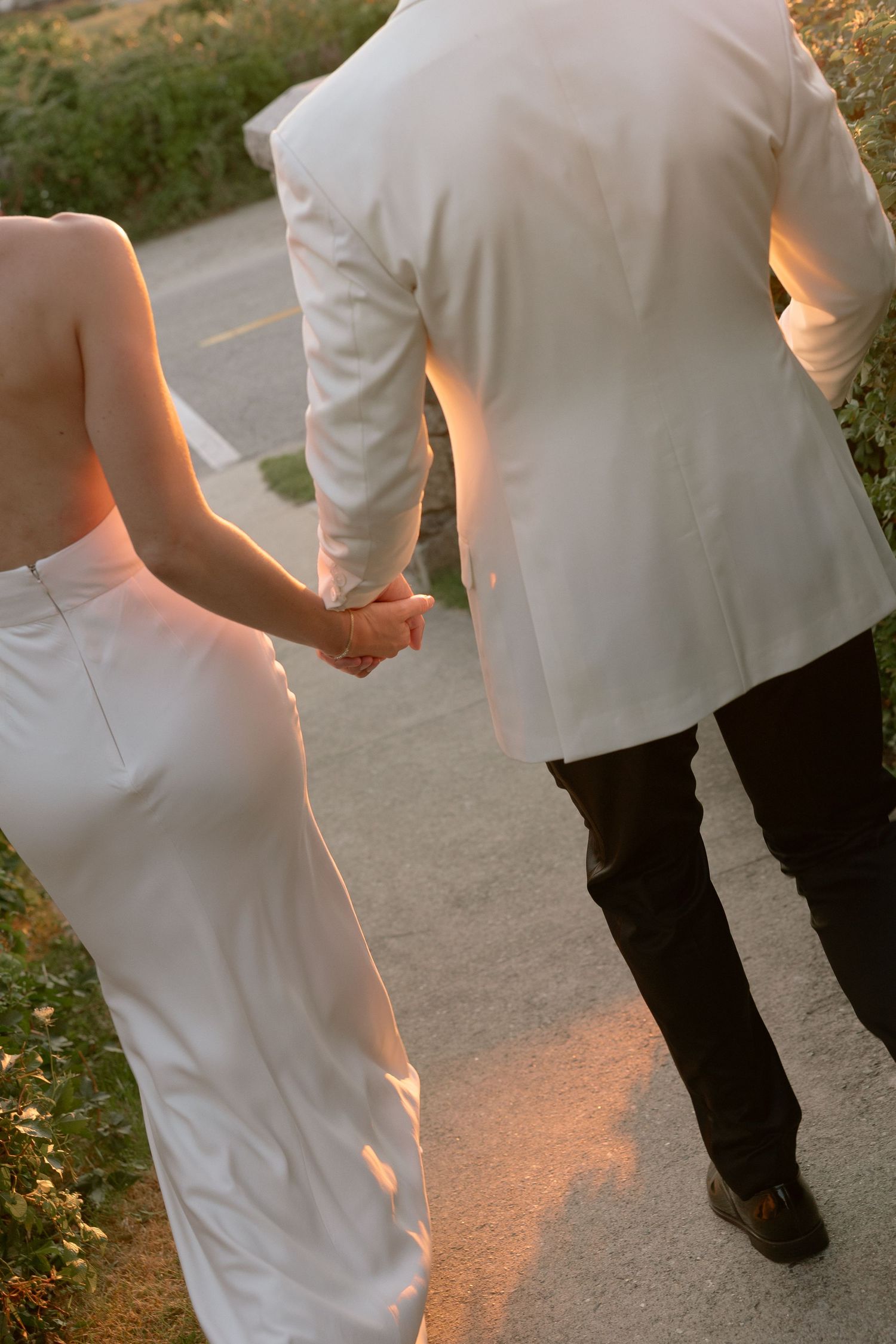 Bride and groom holding hands walking along a sunlit path in elegant white wedding attire at sunset.