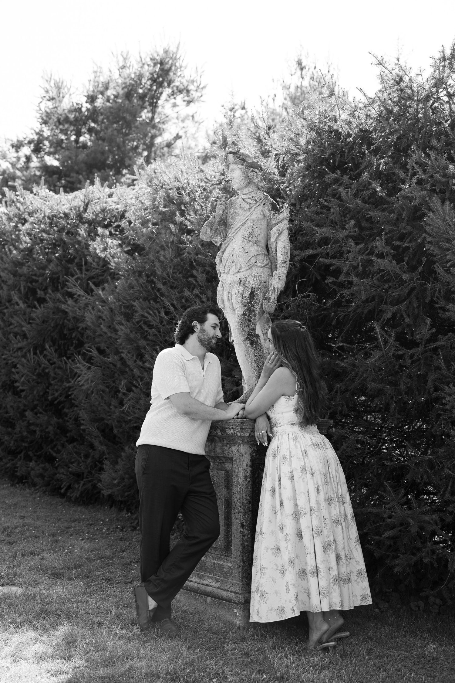 Black and white photo of a couple holding hands near a garden statue surrounded by lush hedges.