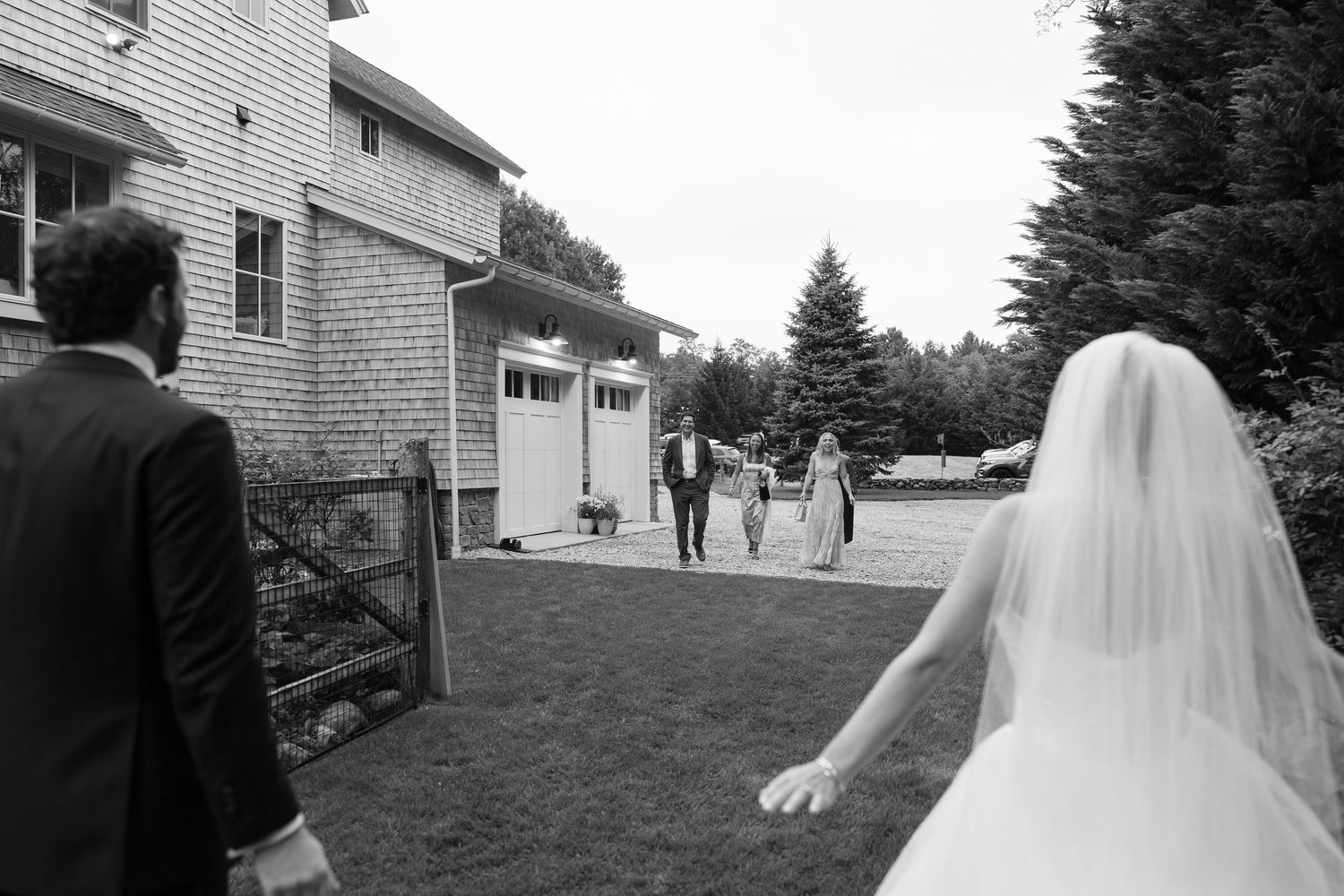 Black and white photo of a bride and groom walking toward wedding guests outside a rustic venue.