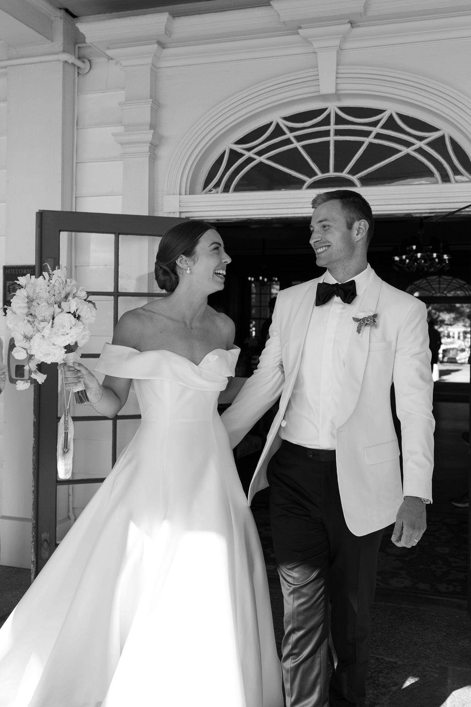 Black and white photo of a smiling bride in off-shoulder gown and groom in white tuxedo walking together at their wedding.
