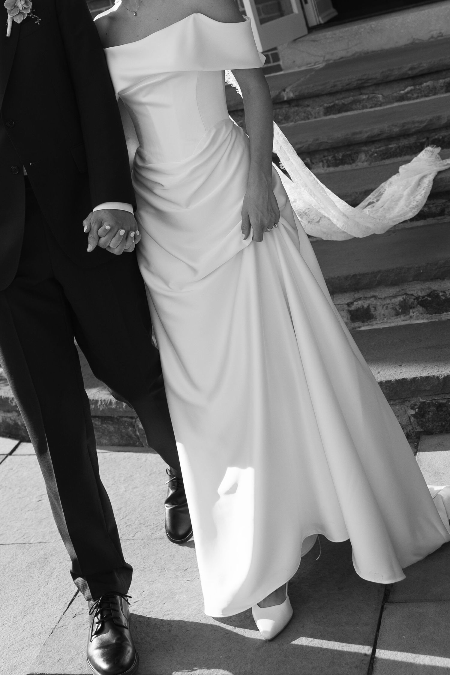 Black and white photo of bride and groom holding hands, bride in elegant off-shoulder wedding gown on stone steps.