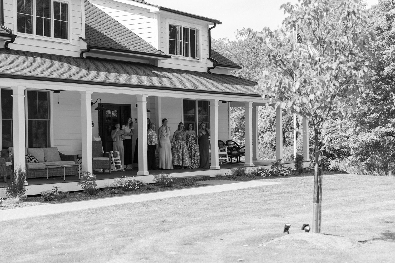 Group of people gathered on a large white farmhouse porch surrounded by lush greenery on a sunny day.
