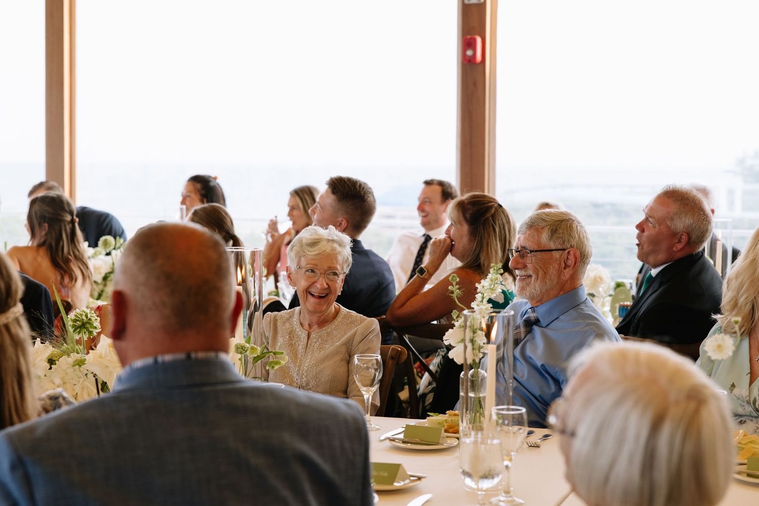 Elegant wedding reception guests seated at round tables with floral centerpieces and scenic views through large windows.
