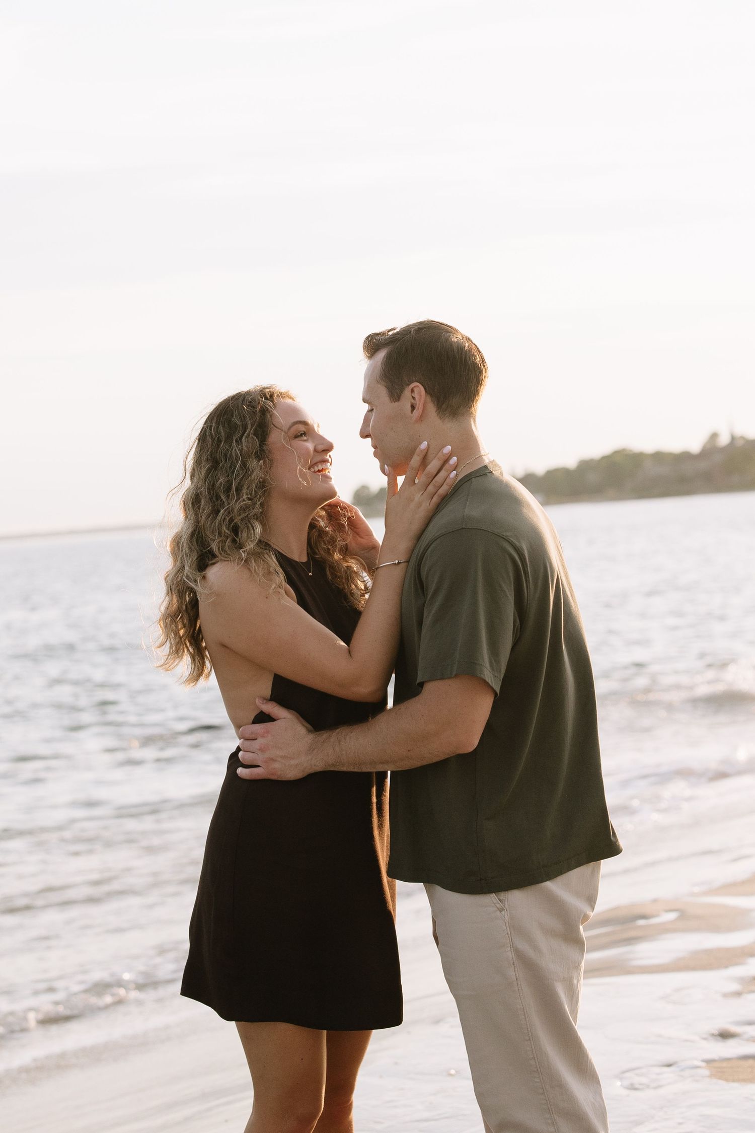 A couple embraces and smiles at each other on a beach at sunset, woman in black dress, man in olive shirt.
