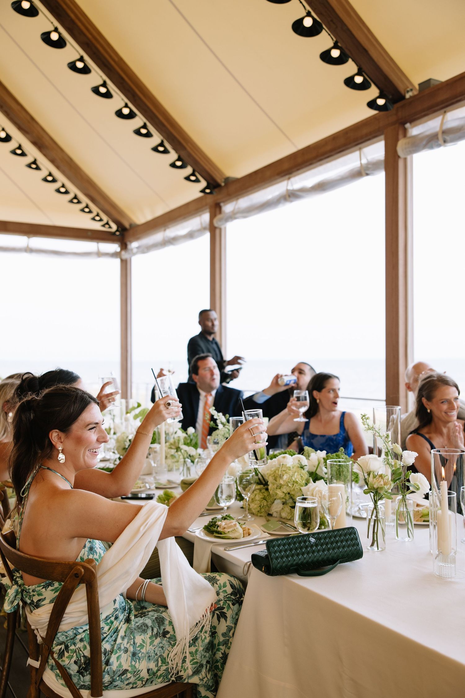 Guests raise glasses in a toast at an elegant outdoor wedding reception under a tent overlooking the ocean.