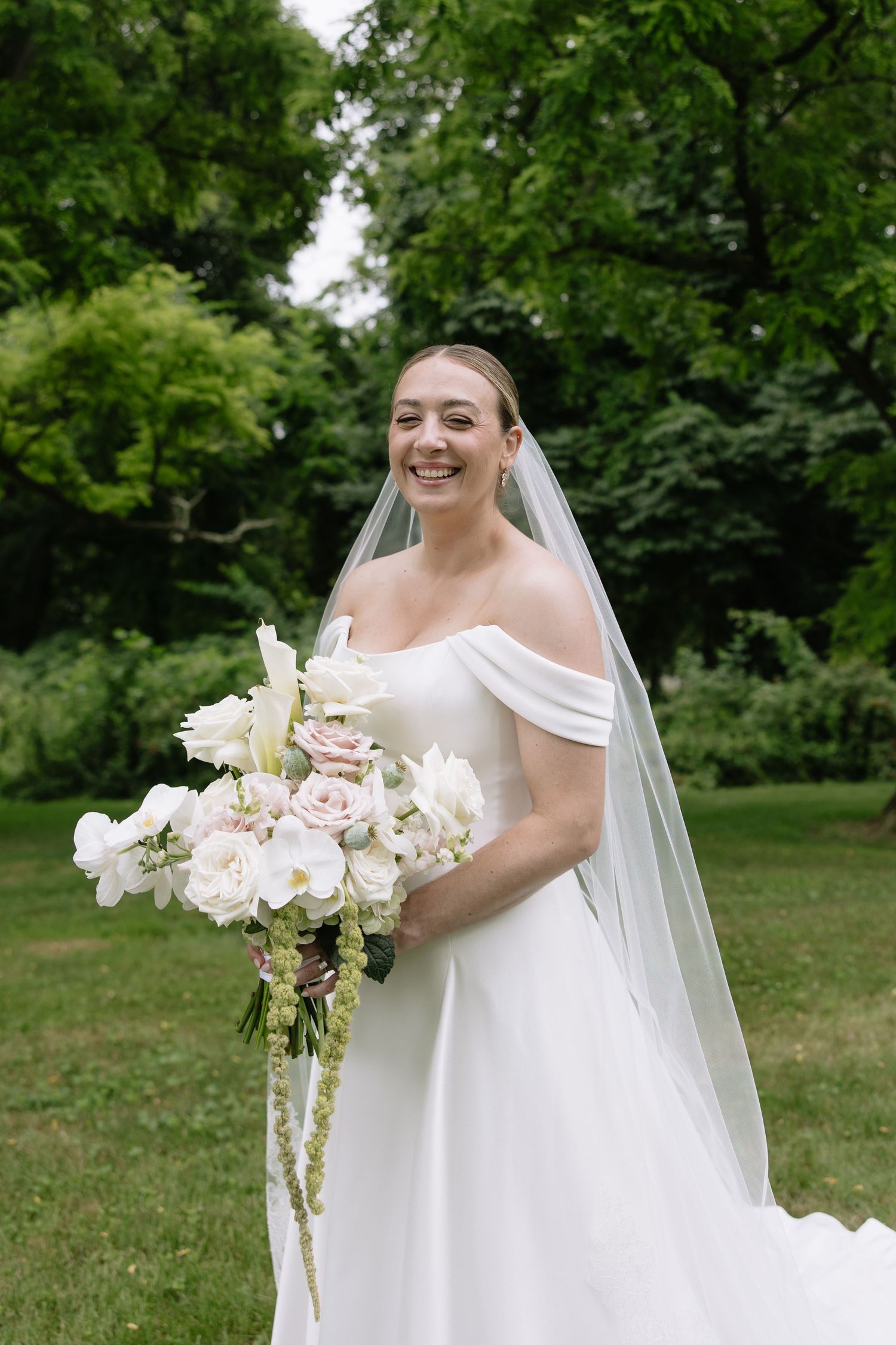 Elegant bride in off-shoulder wedding gown holds luxurious white bouquet in garden setting.