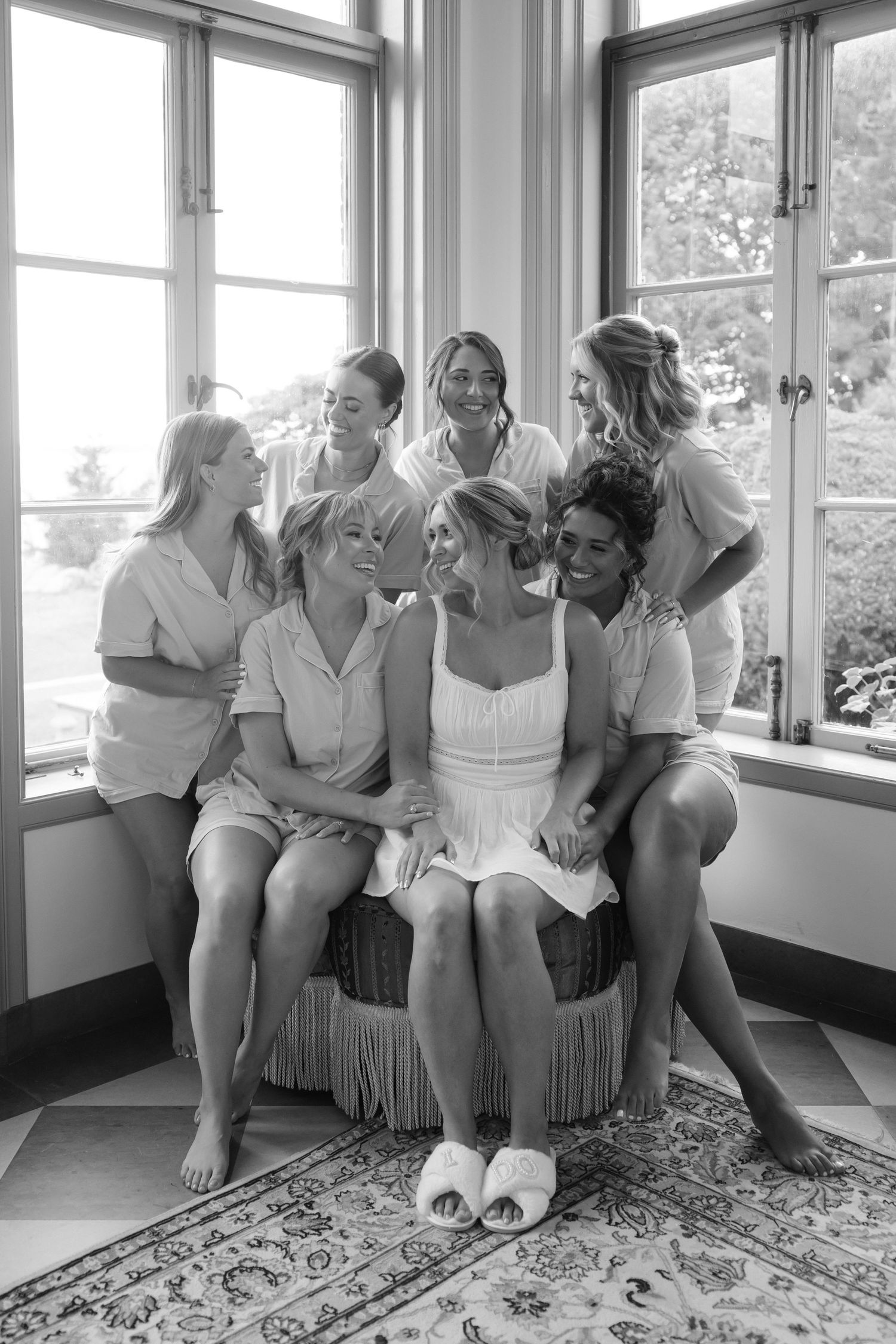 Wedding party in white dresses sits together laughing by bright windows.