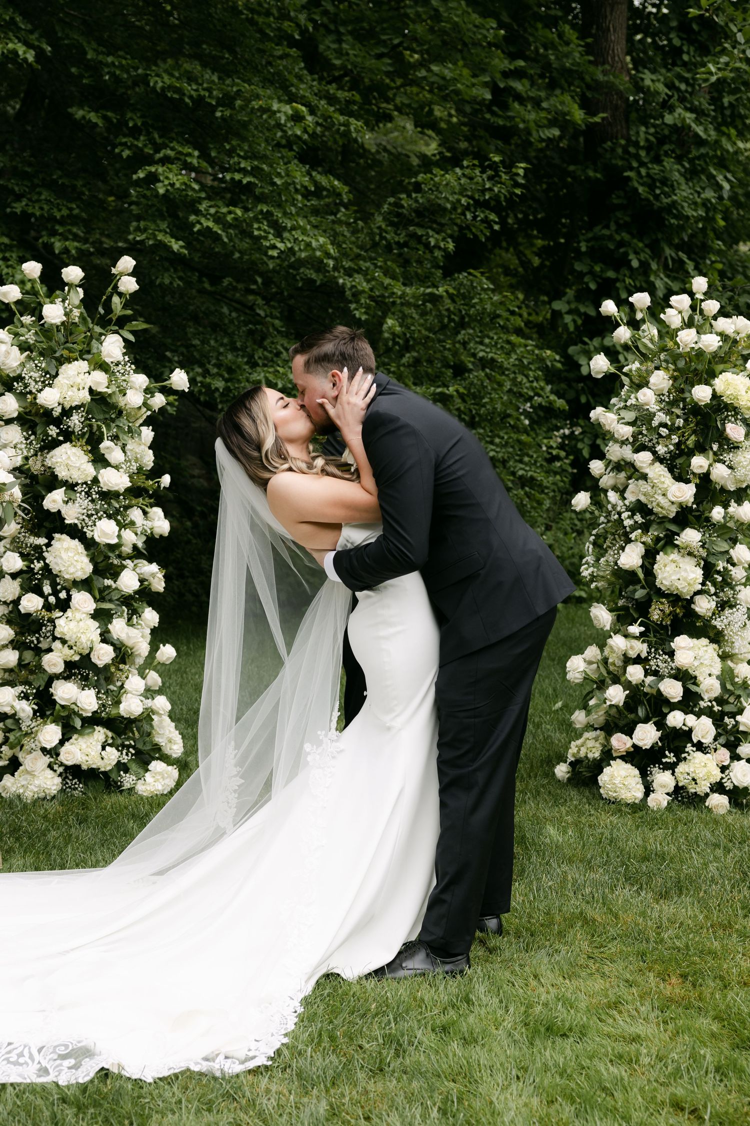 Newlyweds share romantic kiss at outdoor ceremony with white floral arch in background.