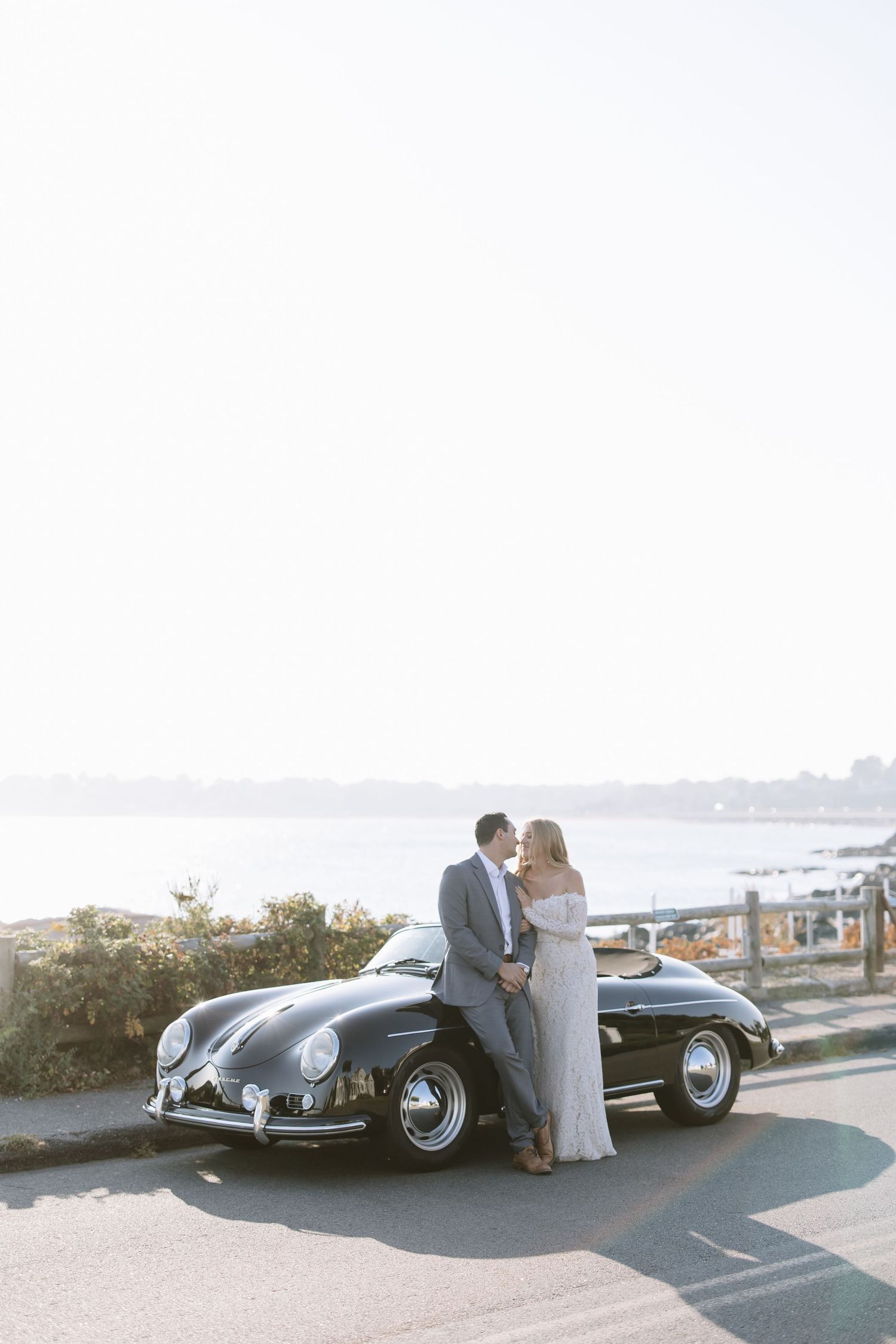 Wedding couple poses with classic vintage cars against a coastal backdrop.