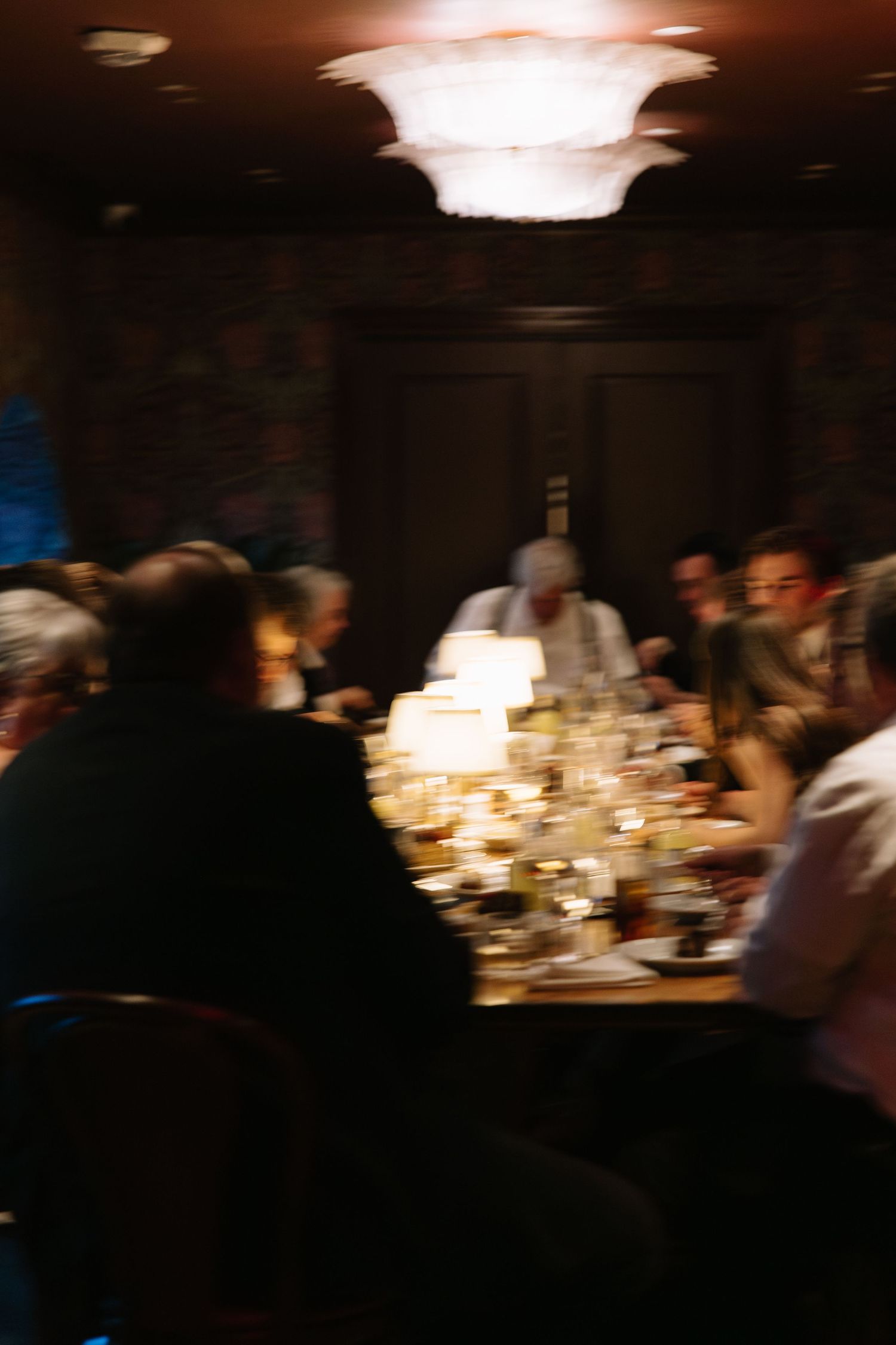 Blurry motion shot of guests dining at a dimly lit restaurant table with warm chandelier lighting overhead.