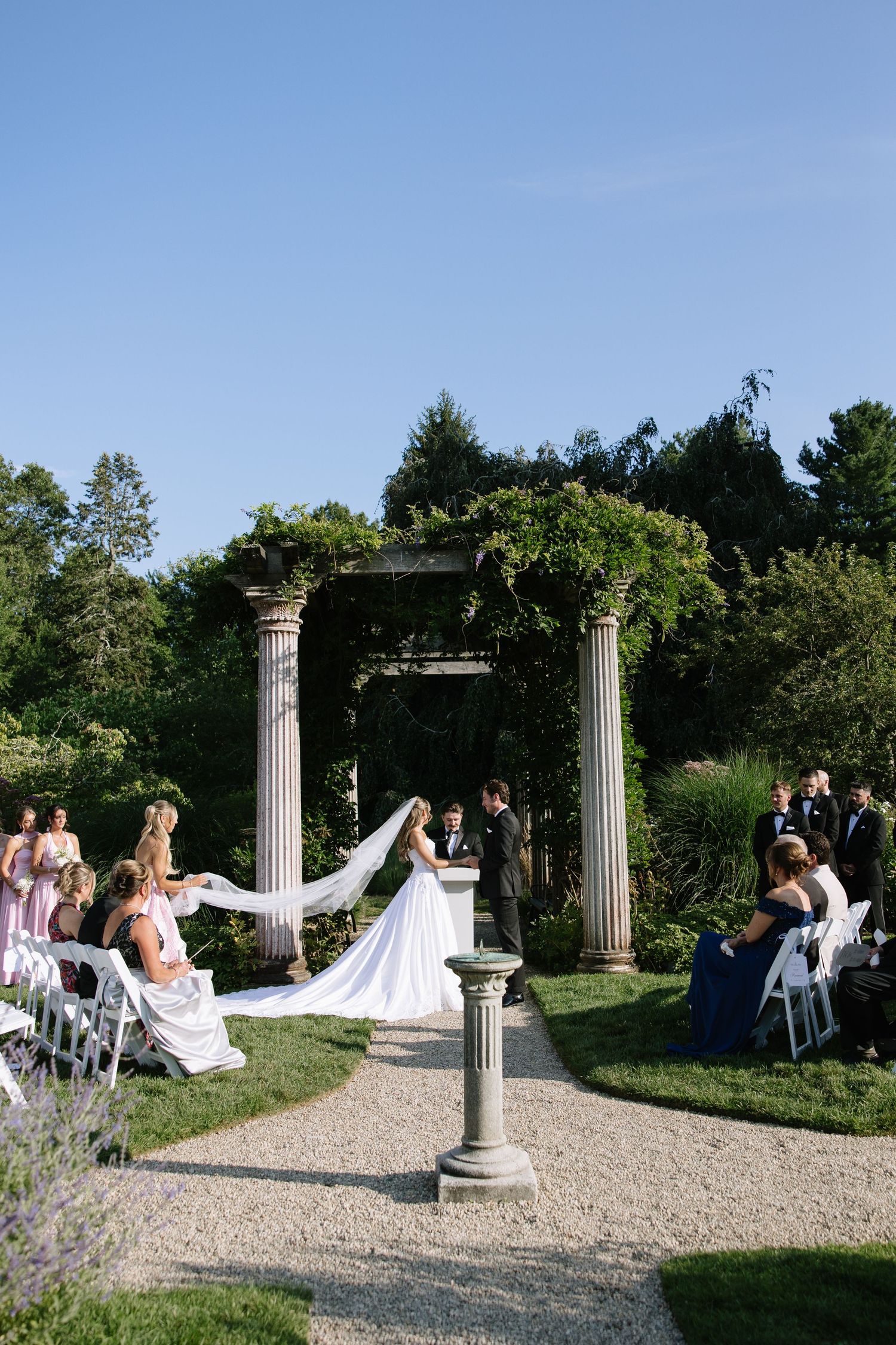 Outdoor garden wedding ceremony under a vine-covered pergola with white chairs and a long bridal veil train.