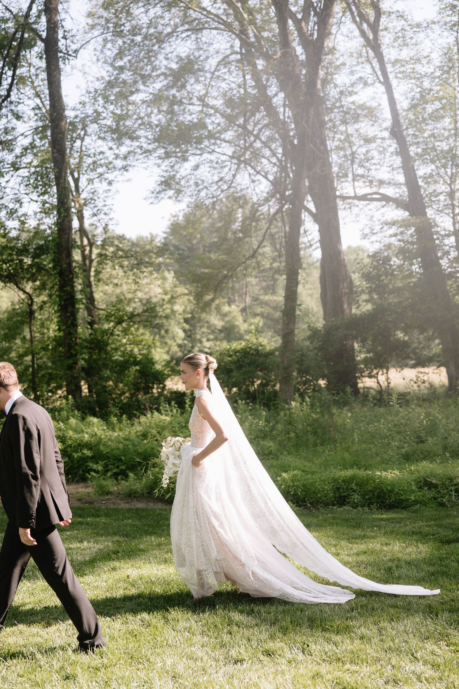 Bride in flowing white gown with long train walks with groom through sunlit garden setting.
