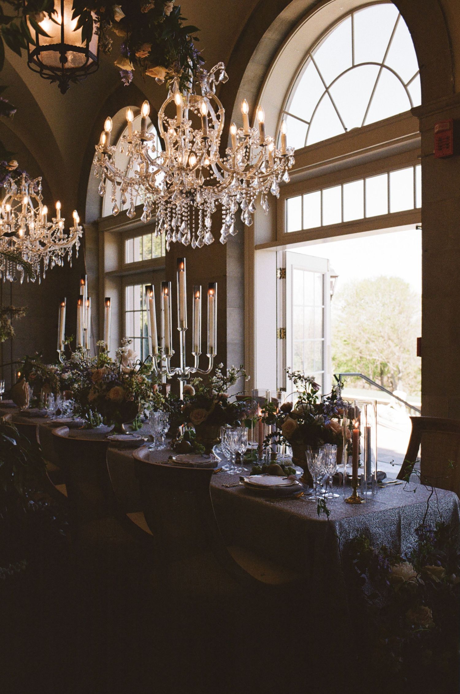 Elegant wedding reception hall with crystal chandeliers, candlelit long tables, floral centerpieces, and arched windows.