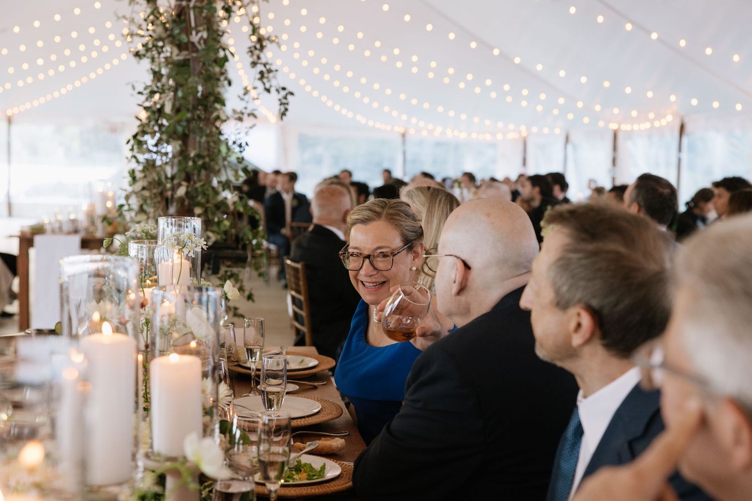 Guests laughing and enjoying dinner under string lights at an elegant tented wedding reception.