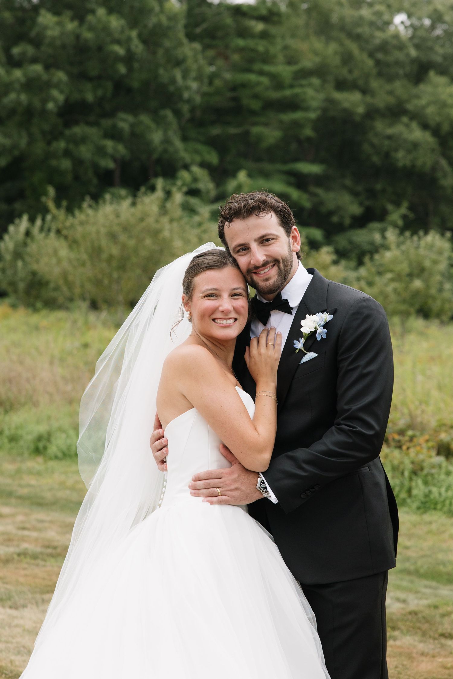 Smiling bride and groom embrace outdoors on their wedding day, she in a white gown with veil, he in a black tuxedo.