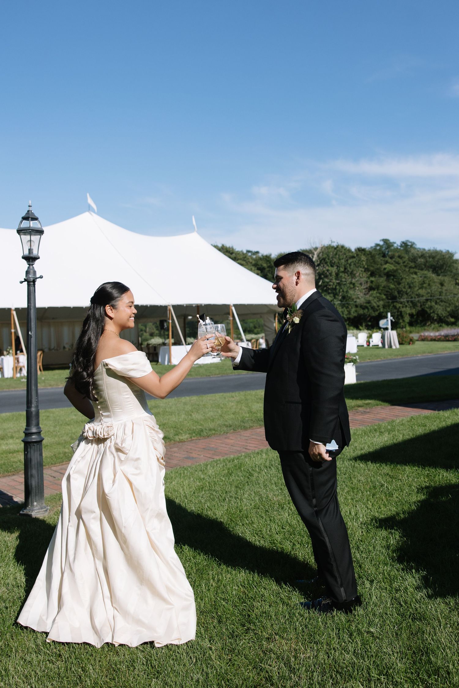 Bride and groom toast drinks on lawn with white reception tent behind.
