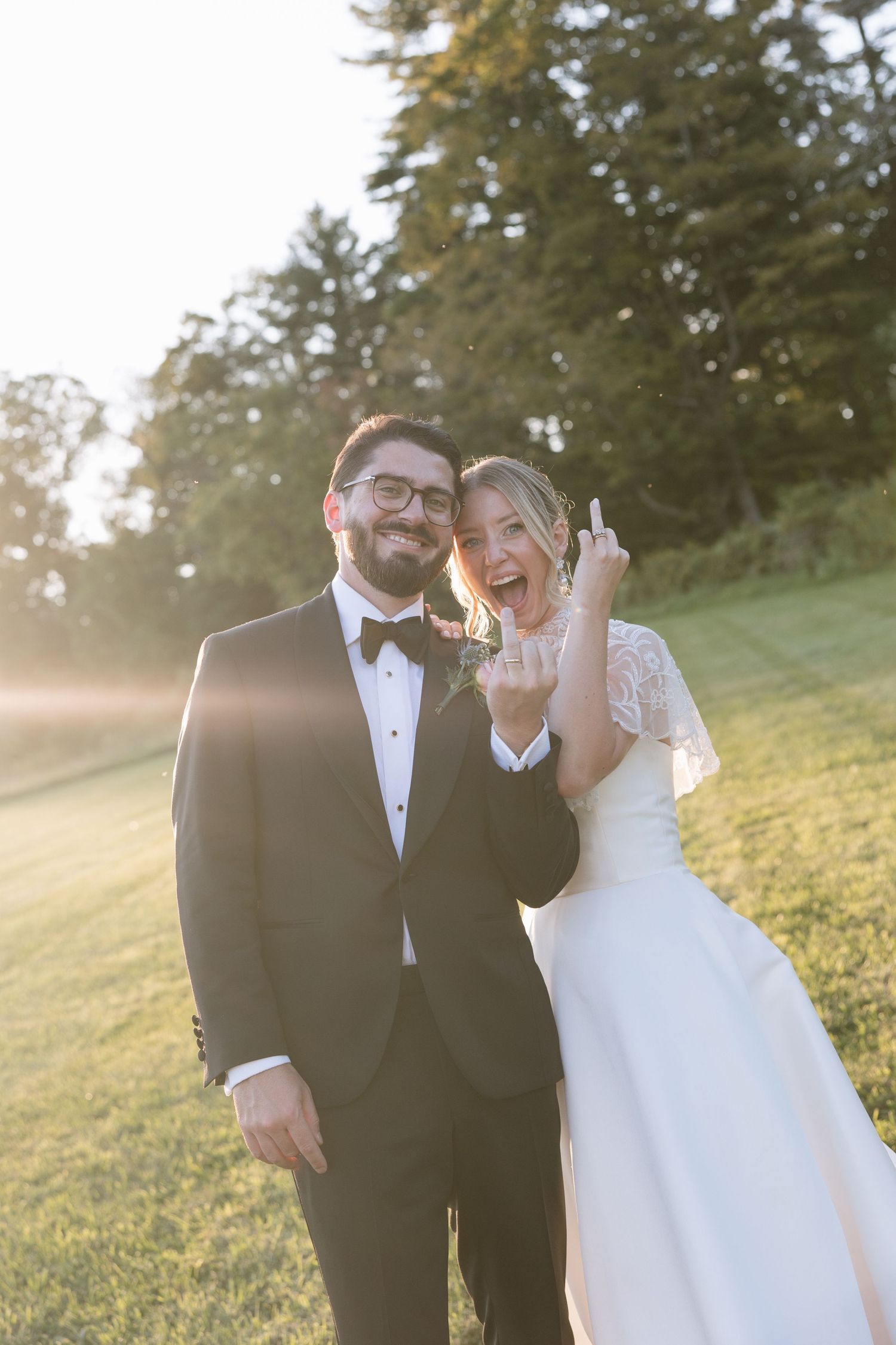 Happy newlyweds posing outdoors at golden hour, bride in white lace gown showing wedding ring, groom in black tuxedo.