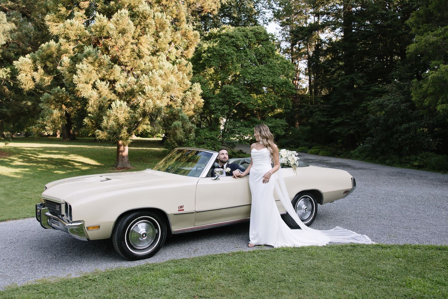 Bride in white gown poses beside a classic cream convertible car with groom seated inside, surrounded by lush green trees.