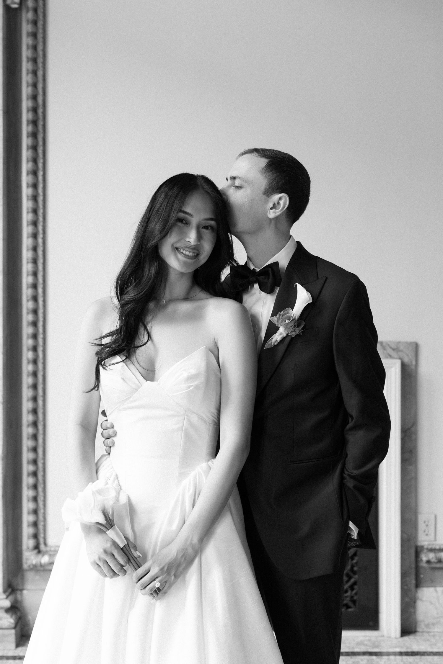 Black and white wedding portrait of smiling bride in strapless gown as groom in tuxedo kisses her cheek.